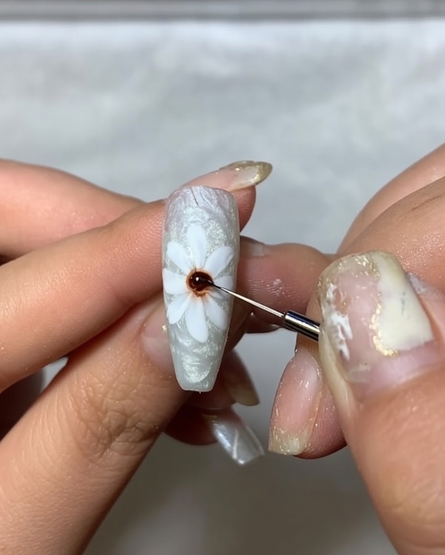 Un primo piano di ununghia con petali bianchi dipinti, usando un pennello fine per applicare un punto di gel marrone scuro al centro.
