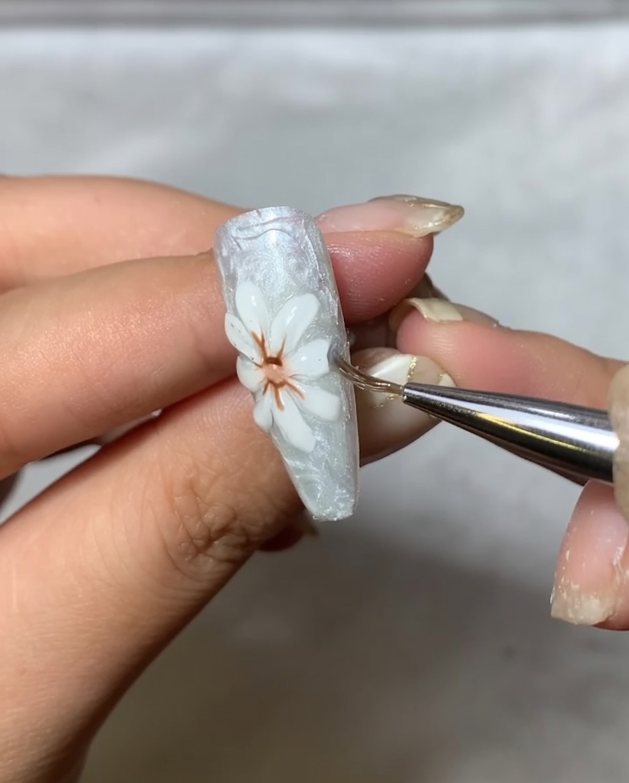 Un pennello applicatore che stende delicatamente polvere a specchio metallica sui petali bianchi rialzati della nail art.