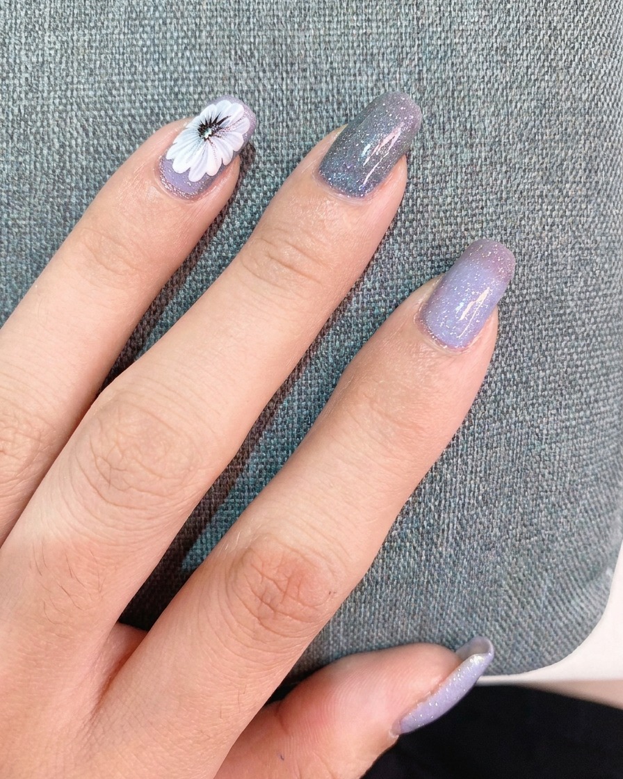Una mano che applica uno strato sottile di top coat sopra una decorazione finita che mostra una margherita bianca e un piccolo adesivo con testo.