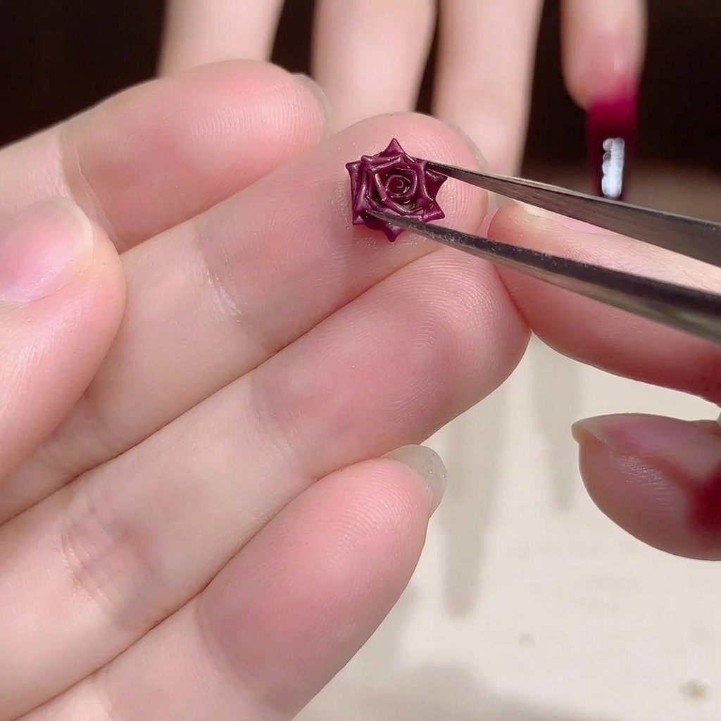 Primo piano di una pinzetta metallica che regge un piccolo e intricato charm per nail art a forma di rosa essiccata rosso scuro.