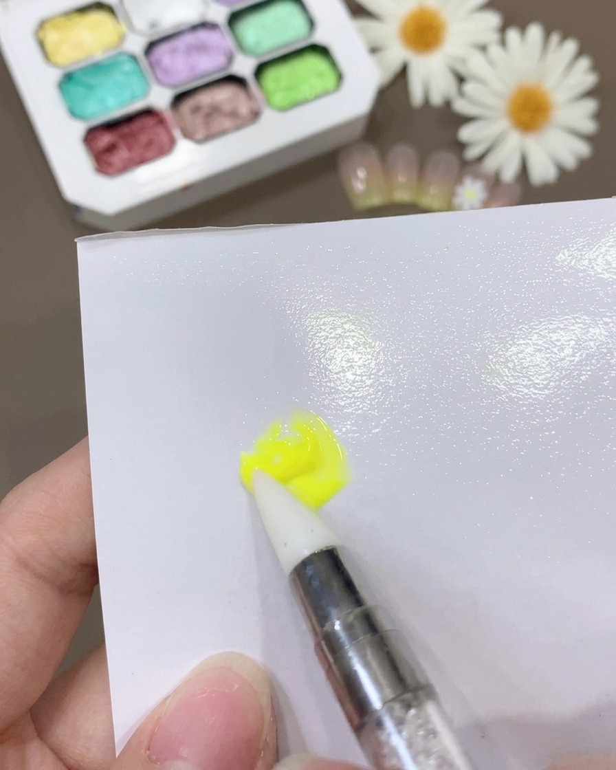 Un pennello che miscela gel giallo e base coat su una superficie bianca per creare una miscela sottile e coerente.