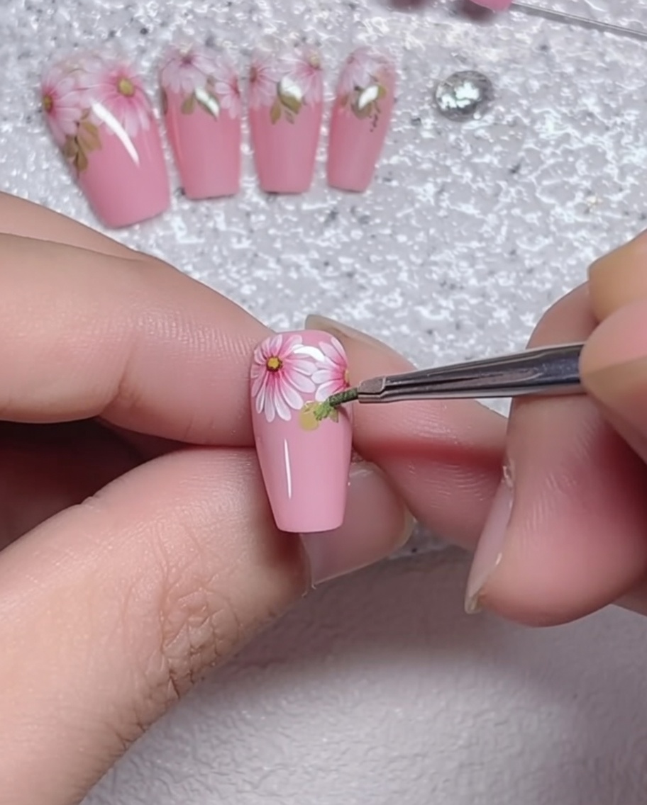 Primo piano di un pennello a punta sottile che aggiunge piccoli dettagli di foglie verdi alla base di una margherita su ununghia rosa.