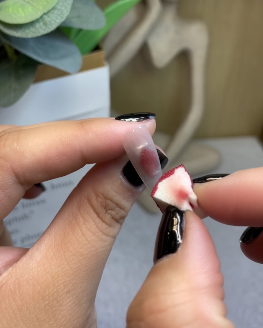 Una spugnetta per maquillage che tampona del gel rosso vino scuro al centro della punta di ununghia trasparente.