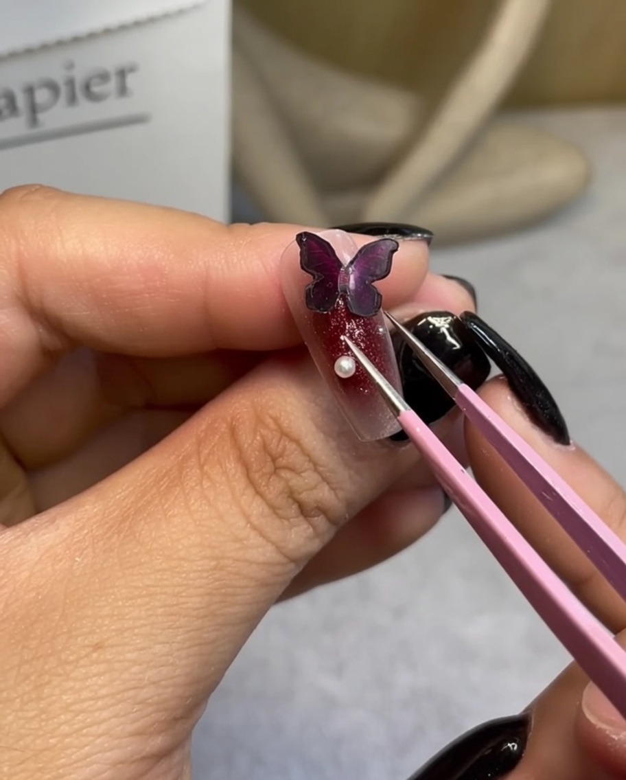 Un paio di pinzette di precisione che posizionano una piccola perla bianca accanto a un charm a farfalla scuro su ununghia curata.