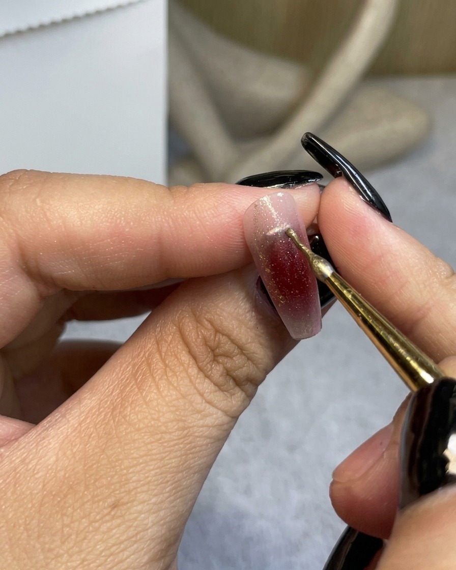 Un pennello a punta fine che applica una sottile linea metallica dorata vicino alle cuticole di ununghia sfumata color rosso vino.