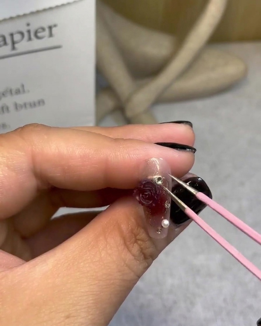 Una mano che utilizza le pinzette per aggiungere minuscole perle attorno a una rosa 3D su un design di unghie completato.
