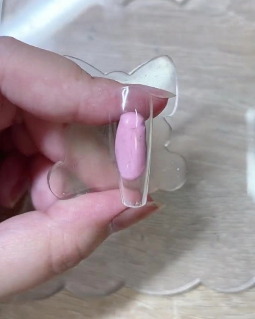 Un primo piano di un pennello a punta fine che disegna delicati contorni di rose bianche su una punta di unghia rosa nuda e traslucida.
