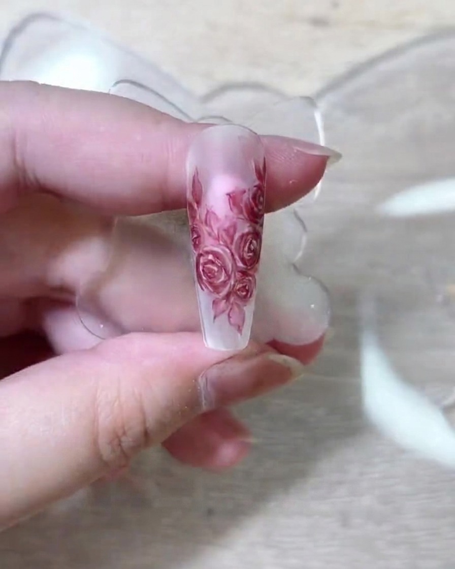 Un pennello fine per nail art che deposita pigmento rosso profondo sui petali di rosa stratificati su unestensione per unghie nude.
