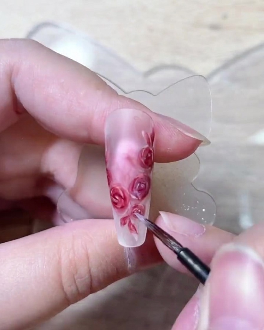 Un primo piano di un pennello per unghie fine che posiziona delicatamente punti luce bianchi sul bordo dei petali di rosa dipinti.