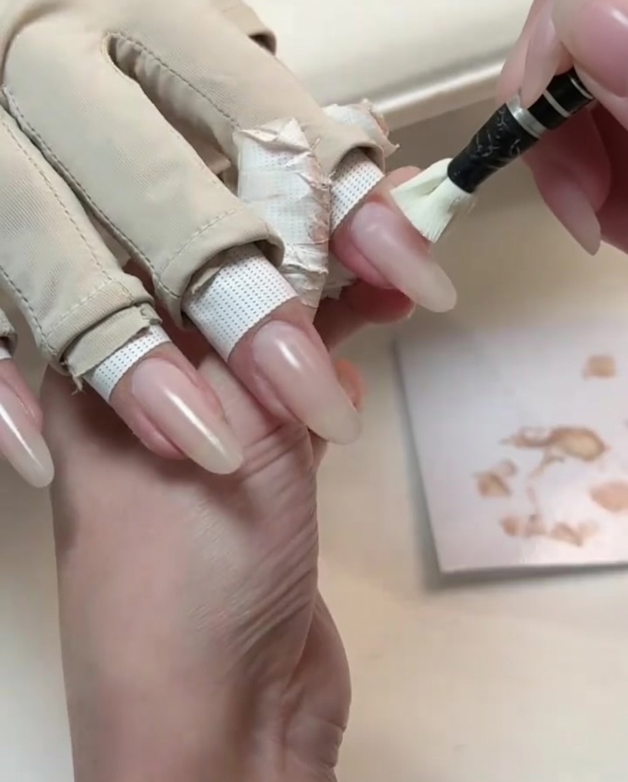 Un piccolo pennello applica un gel di base color nude su ununghia dalla forma naturale lunga.