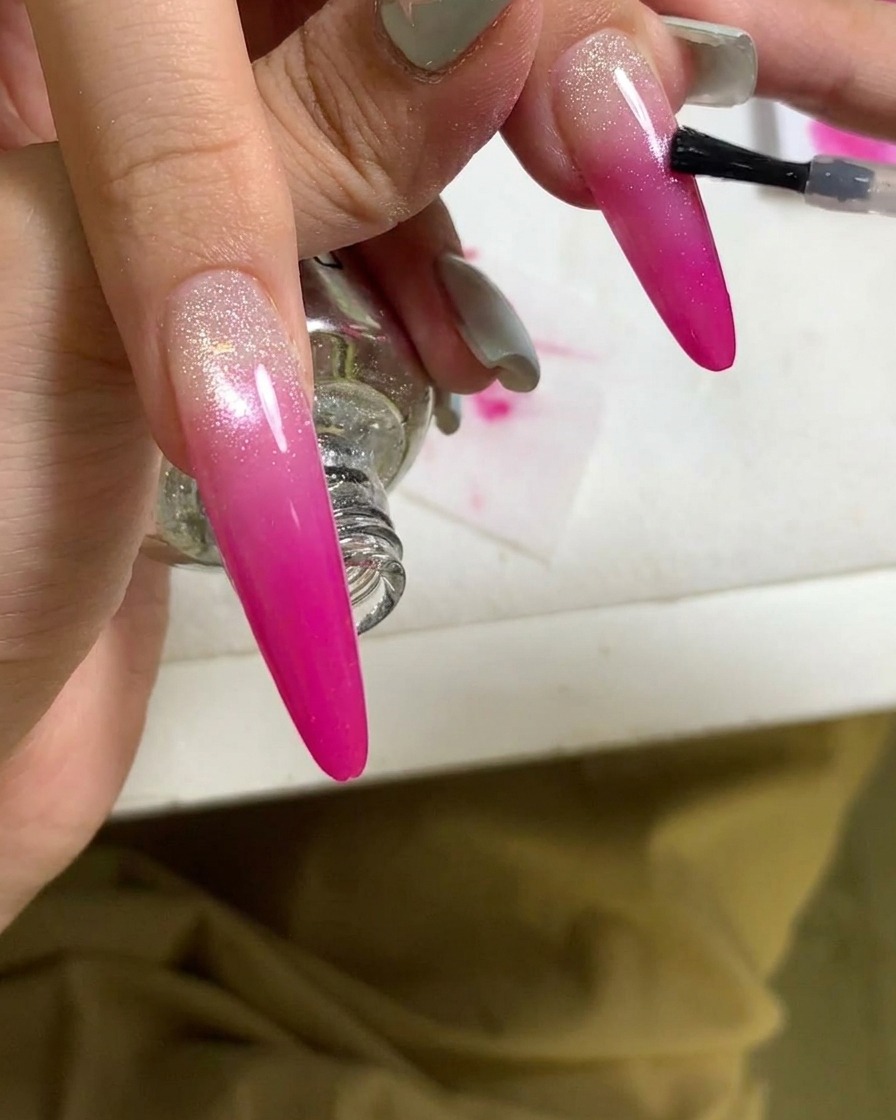 Primo piano di un pennello che applica un top coat su ununghia sfumata rosa finita.