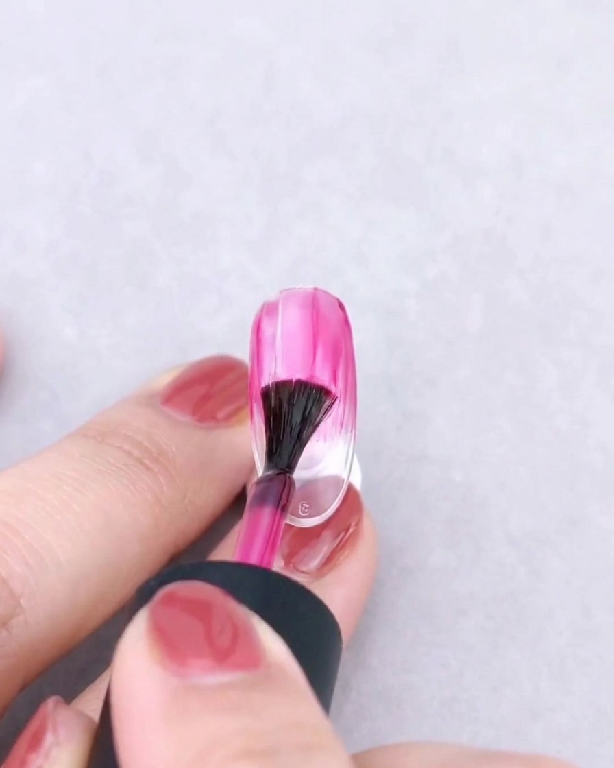 Un pennello a ventaglio che sfufa delicatamente un colore in gel rosa verso il basso su ununghia trasparente per creare un effetto sfumato.