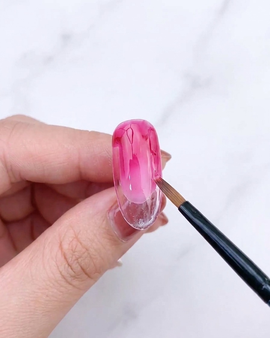 Un pennello a ventaglio che sfuma il secondo strato di gel rosa su ununghia, creando una sfumatura raffinata.