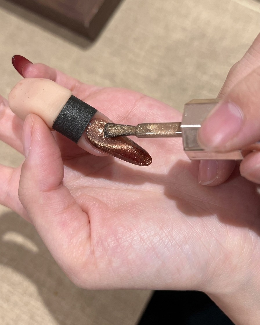 Un pennello per unghie che applica un gel cat eye dorato brillante sopra lunghia con sfumatura rossa.