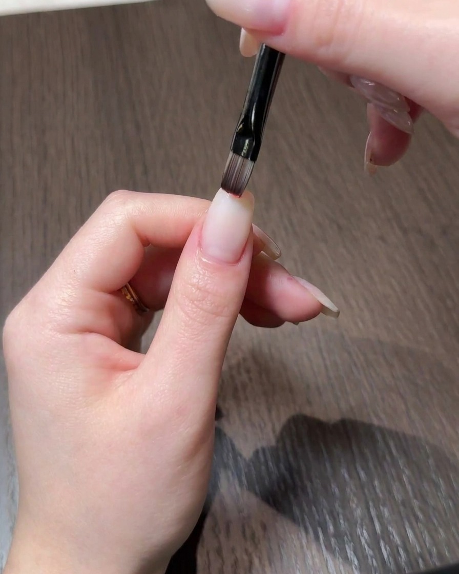 Primo piano di una mano che applica lo smalto gel rosso sulla punta di ununghia dallaspetto naturale usando un pennello sottile.
