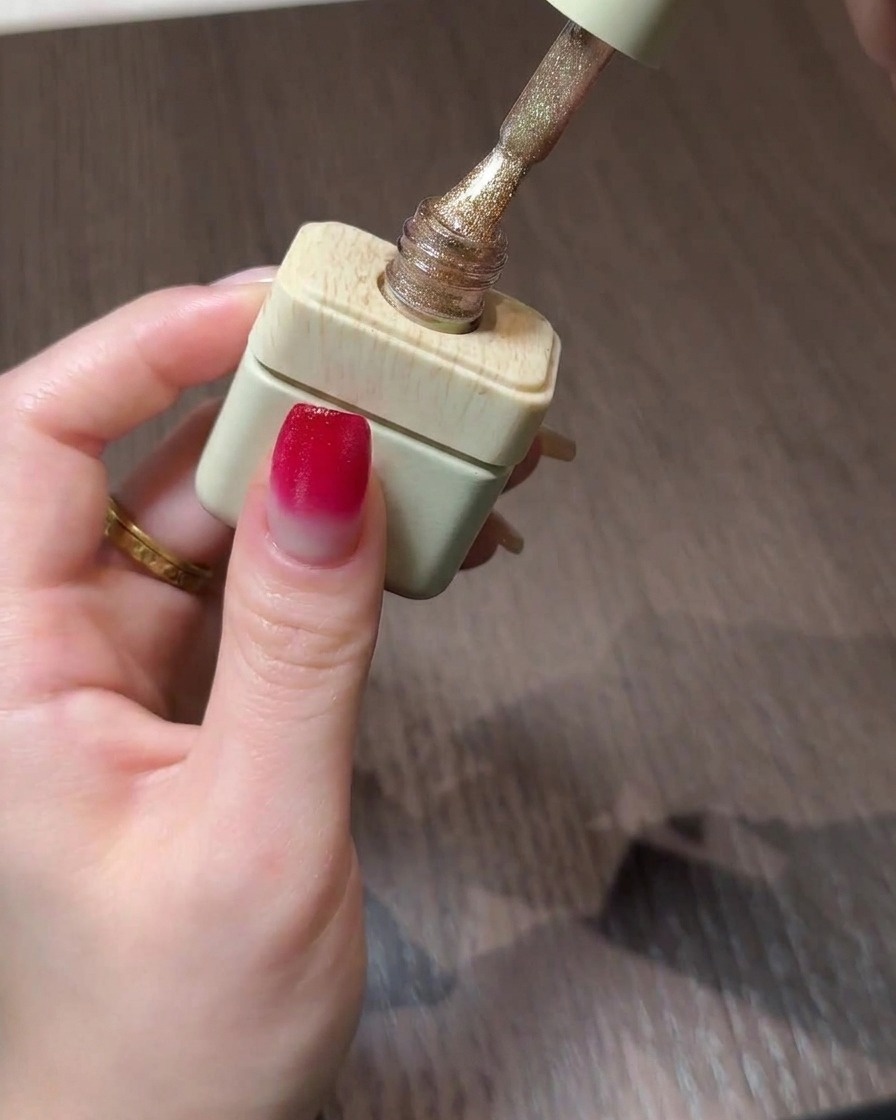 Un flacone di gel polish cat eye dorato con il pennello sospeso sopra ununghia sfumata.
