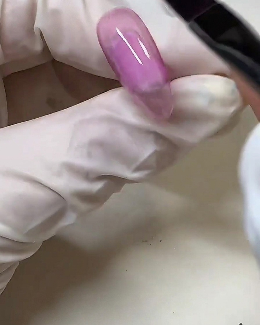Una mano con guanto utilizza un pennellino per gel per applicare lo smalto gel rosa semitrasparente su una tip trasparente.