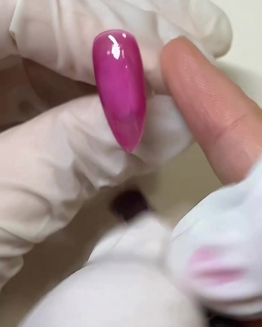 Primo piano di un dito con guanto che applica uno strato sottile di colla solida trasparente sulla superficie dellunghia rosa polimerizzata.