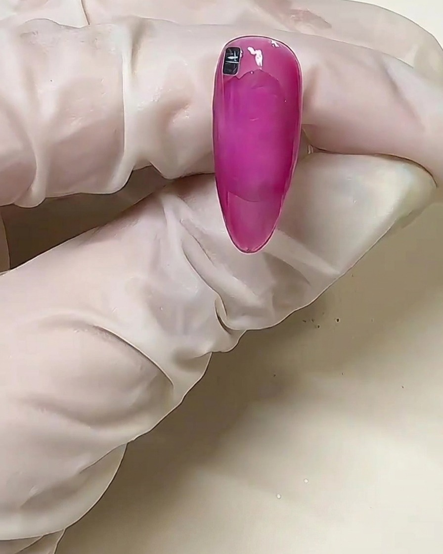 Primo piano di ununghia rosa con un singolo quadratino metallico argentato posizionato vicino alla linea delle cuticole sulla colla solida non polimerizzata.