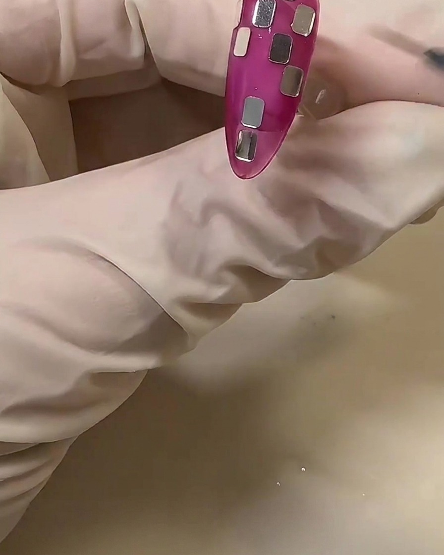Una mano con guanto sistema dei piccoli frammenti di pellicola quadrati argentati su ununghia rosa traslucida per creare un motivo a scacchi.