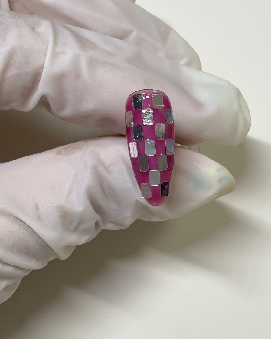 Una mano con guanto tiene ununghia con un motivo a scacchi che è stata rivestita con uno strato di gel rosa velato per uniformare i quadratini argentati.