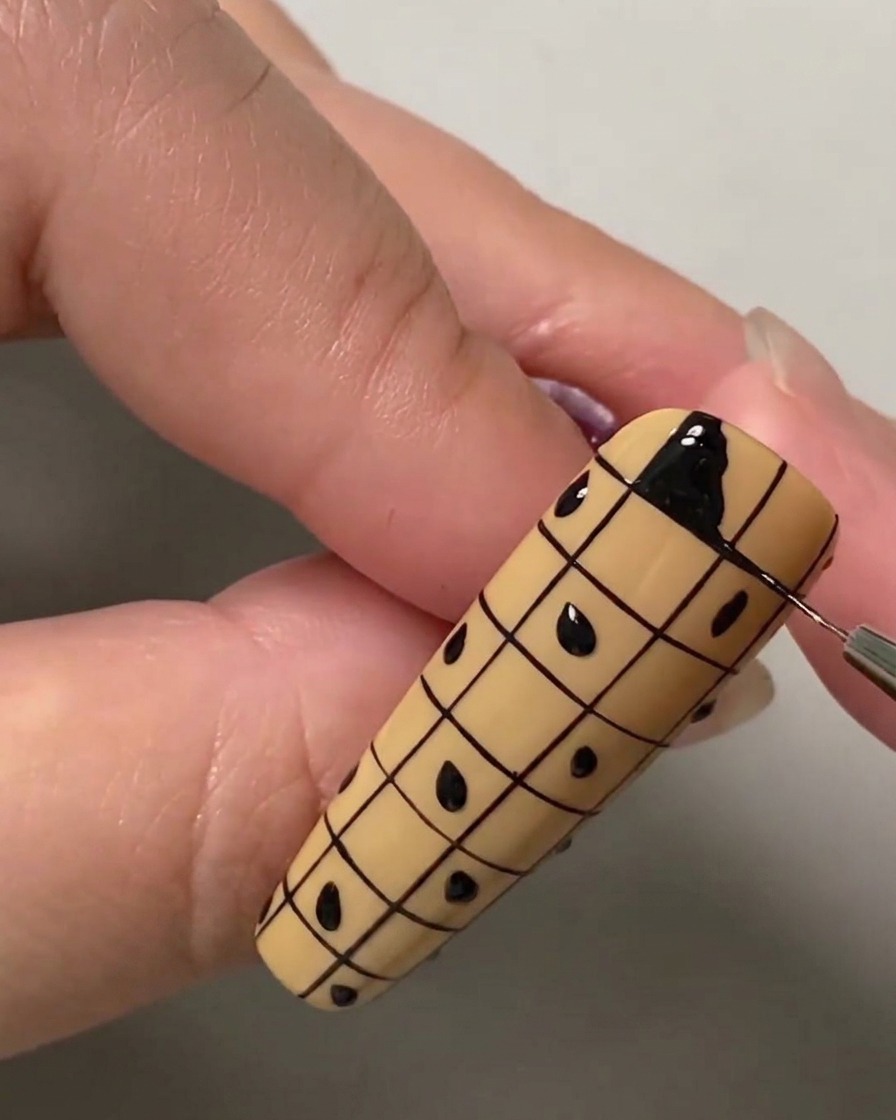 Primo piano di un pennello liner che riempie il quadrato in alto a sinistra di una griglia su ununghia nude usando gel polish nero.