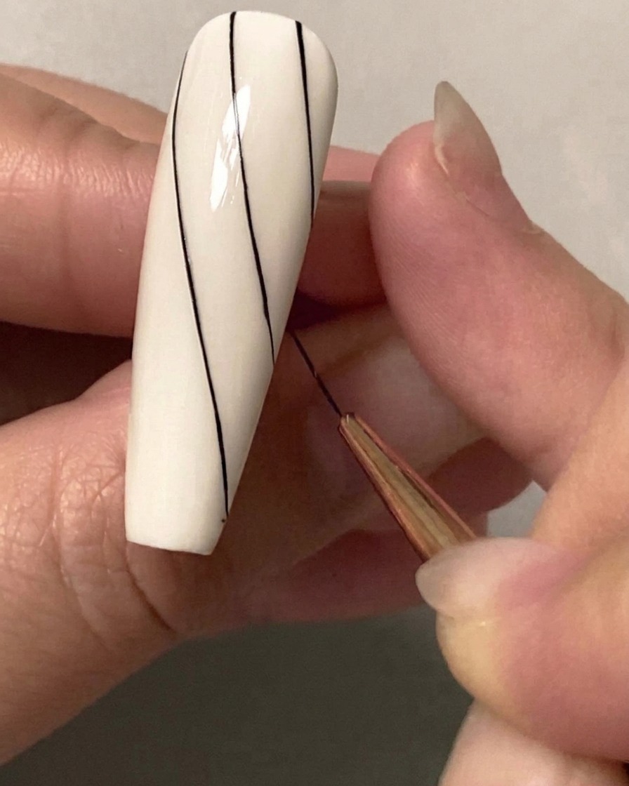 Primo piano di un pennello sottile per nail art che disegna una seconda linea diagonale nera parallela sulla metà superiore di ununghia bianca.