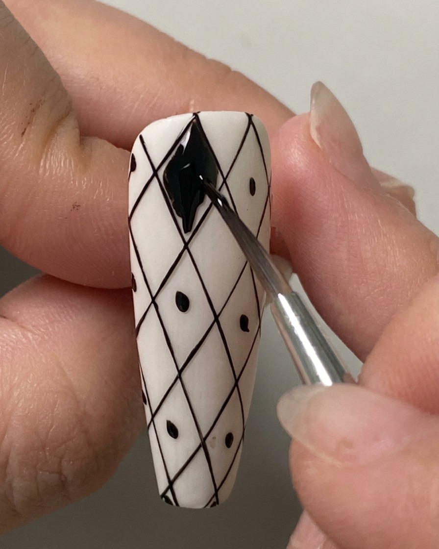 Un pennello liner sottile che riempie con cura la forma a rombo più in alto con gel polish nero su ununghia con griglia bianca pre-disegnata.