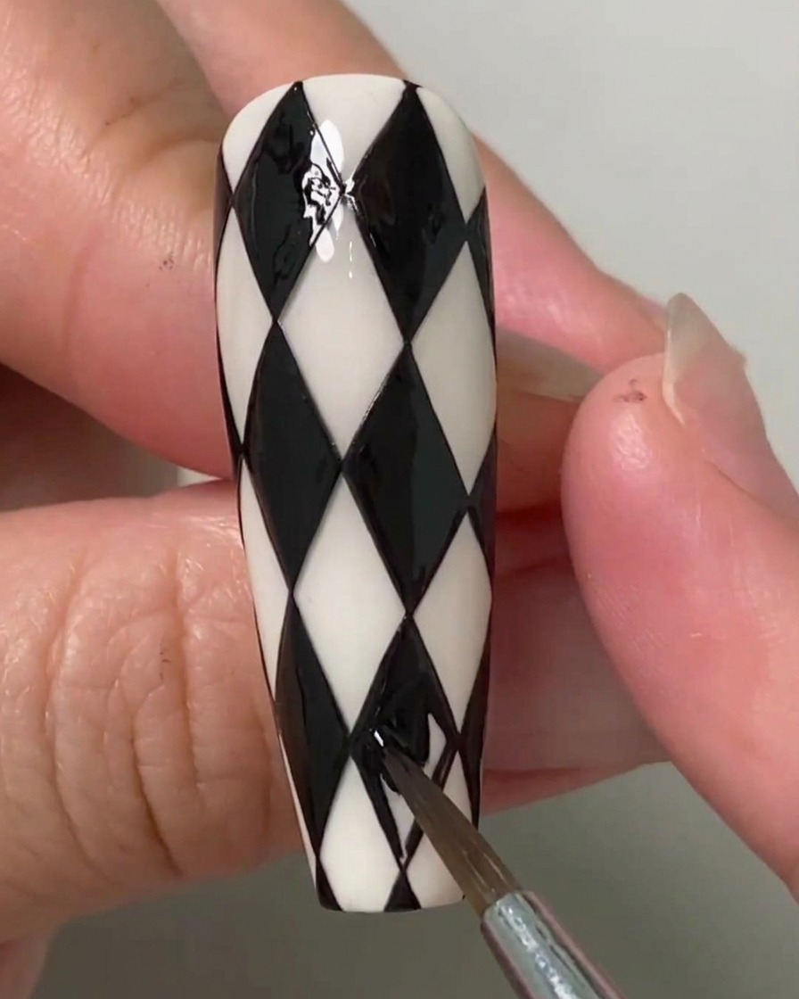 Una nail artist che utilizza un pennello sottile per riempire le ultime forme a rombo rimanenti su ununghia decorata con motivo argyle bianco e nero.