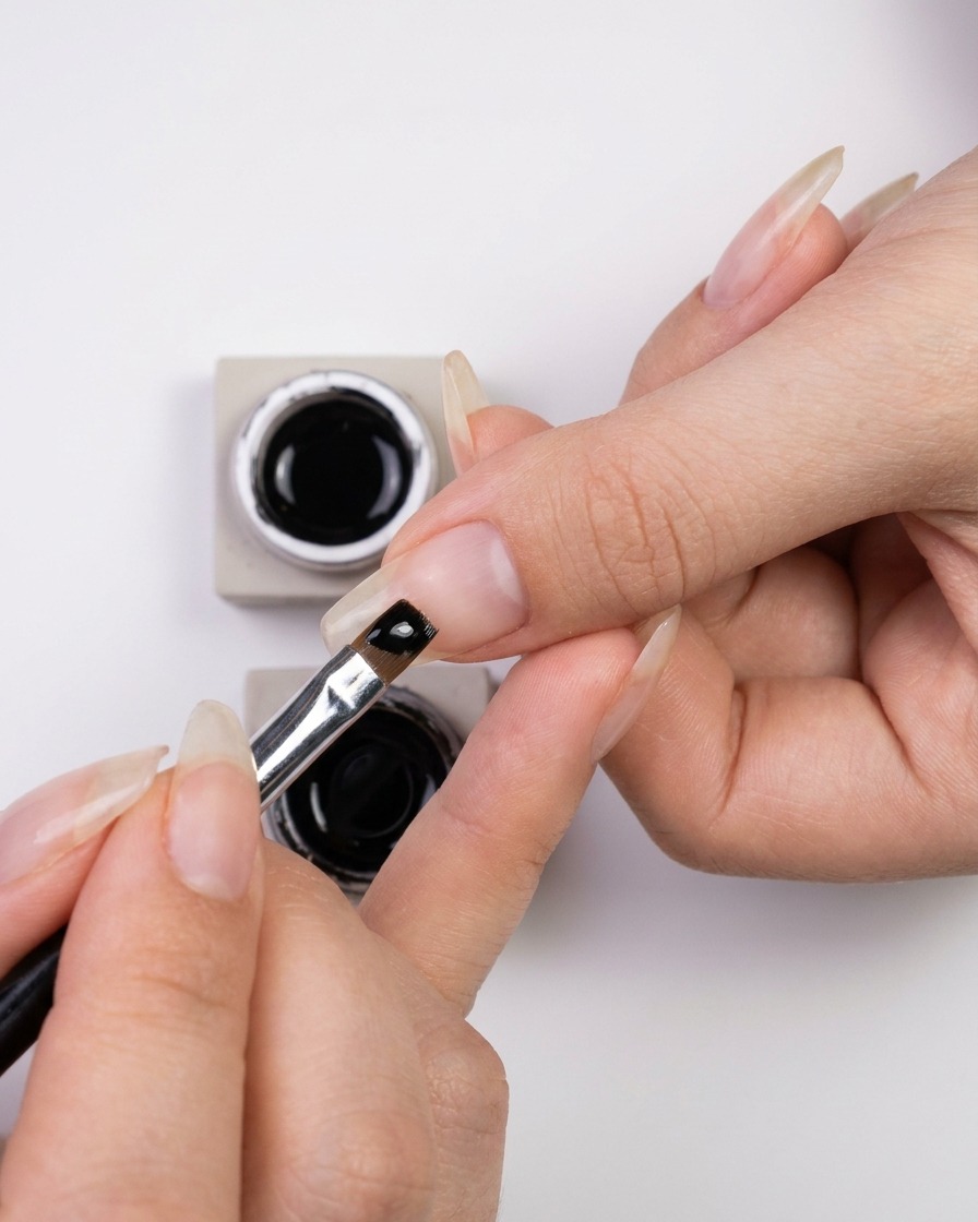 Una mano che intinge un pennello fine per nail art in un piccolo barattolo di smalto gel nero lucido.