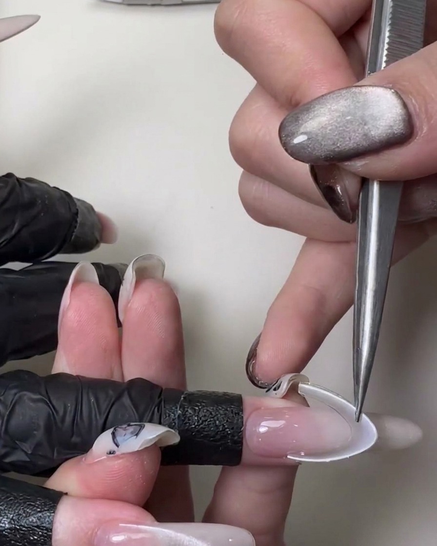 Primo piano di una mano che usa pinzette di metallo per applicare un adesivo curvo bianco per il french su una lunga unghia a stiletto rosa traslucido.