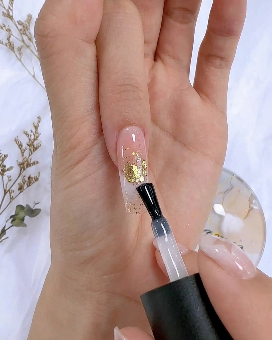 Un pennellino applica un top coat trasparente su ununghia decorata con accenti in foglia doro.