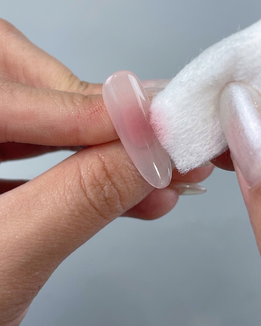 Una mano che usa un mattoncino in spugna bianca per tamponare lo smalto gel rosa al centro di una tip a mandorla trasparente.