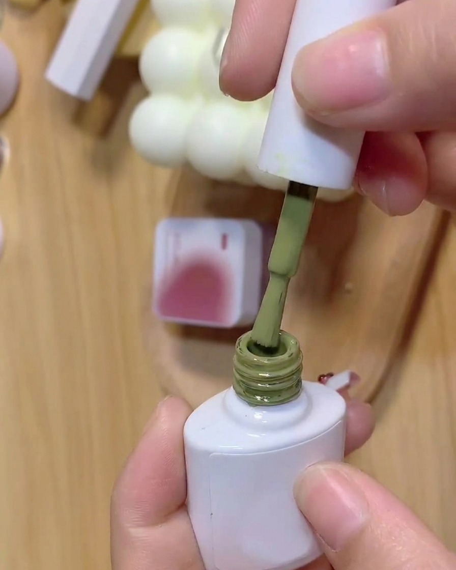 Un pennello si intinge in un flacone di smalto gel verde con sullo sfondo un barattolo di gel rosa.