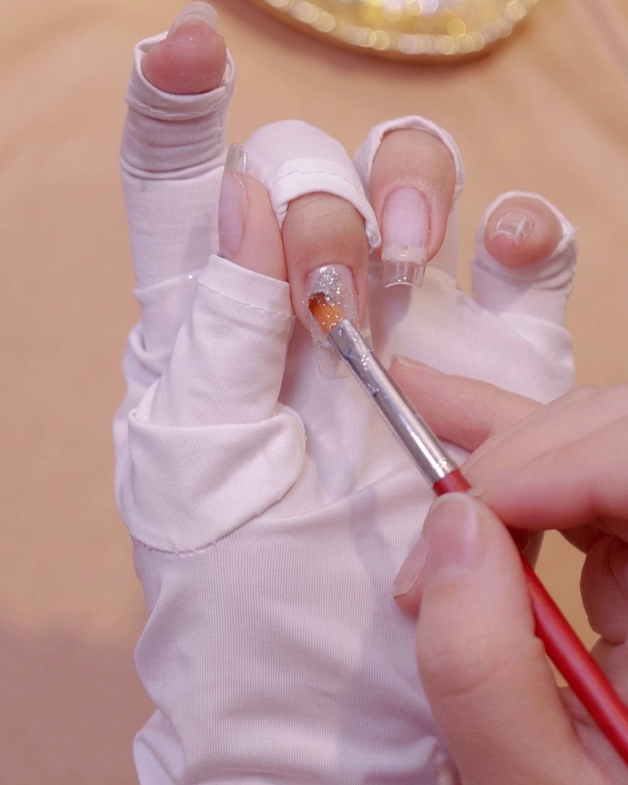 Un pennello fine che applica gel glitterato argento vicino alla base di unestensione per unghie trasparente su una mano guantata.