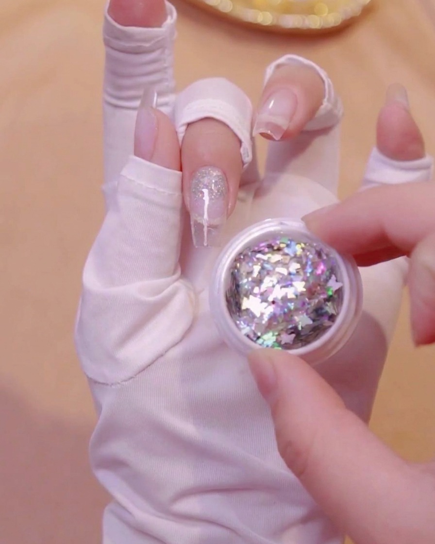 Una mano che tiene un piccolo barattolo aperto pieno di paillettes glitterate olografiche a forma di farfalla.