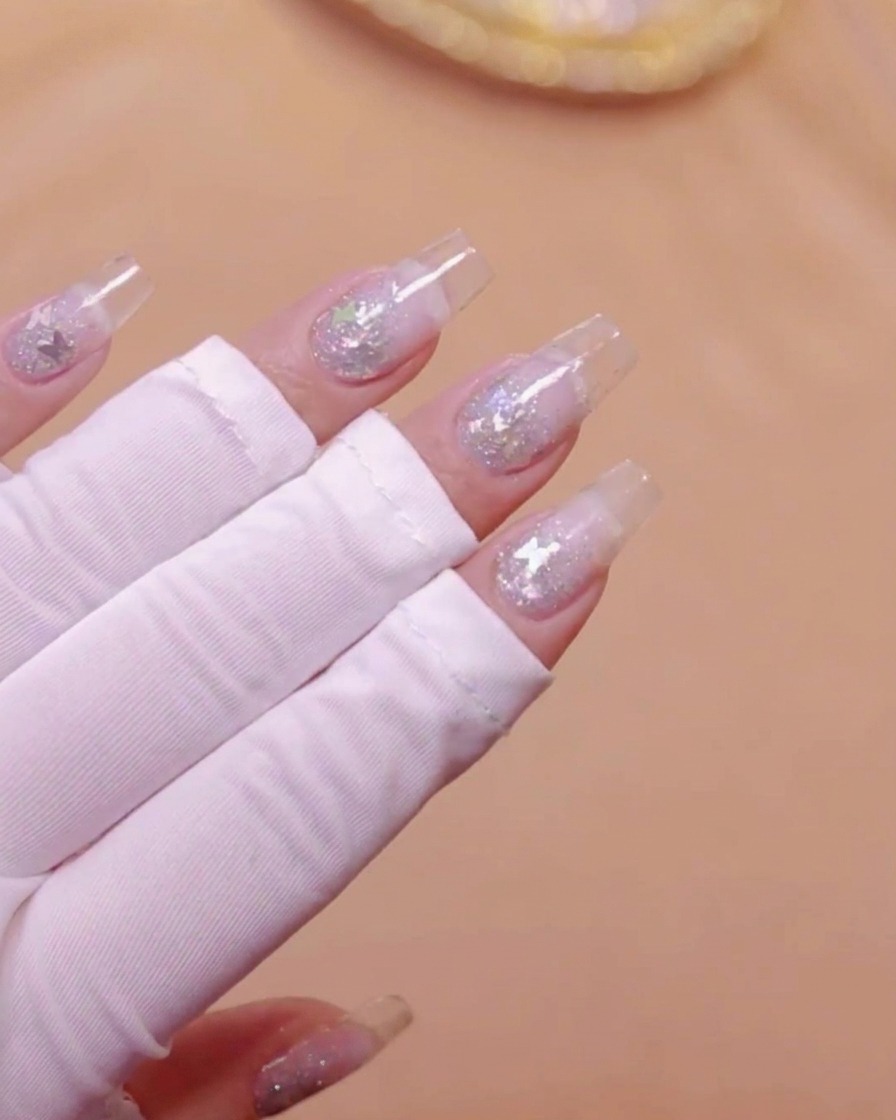 Una mano con estensioni per unghie trasparenti e glitter a farfalla incorporati posizionata sotto una lampada UV.