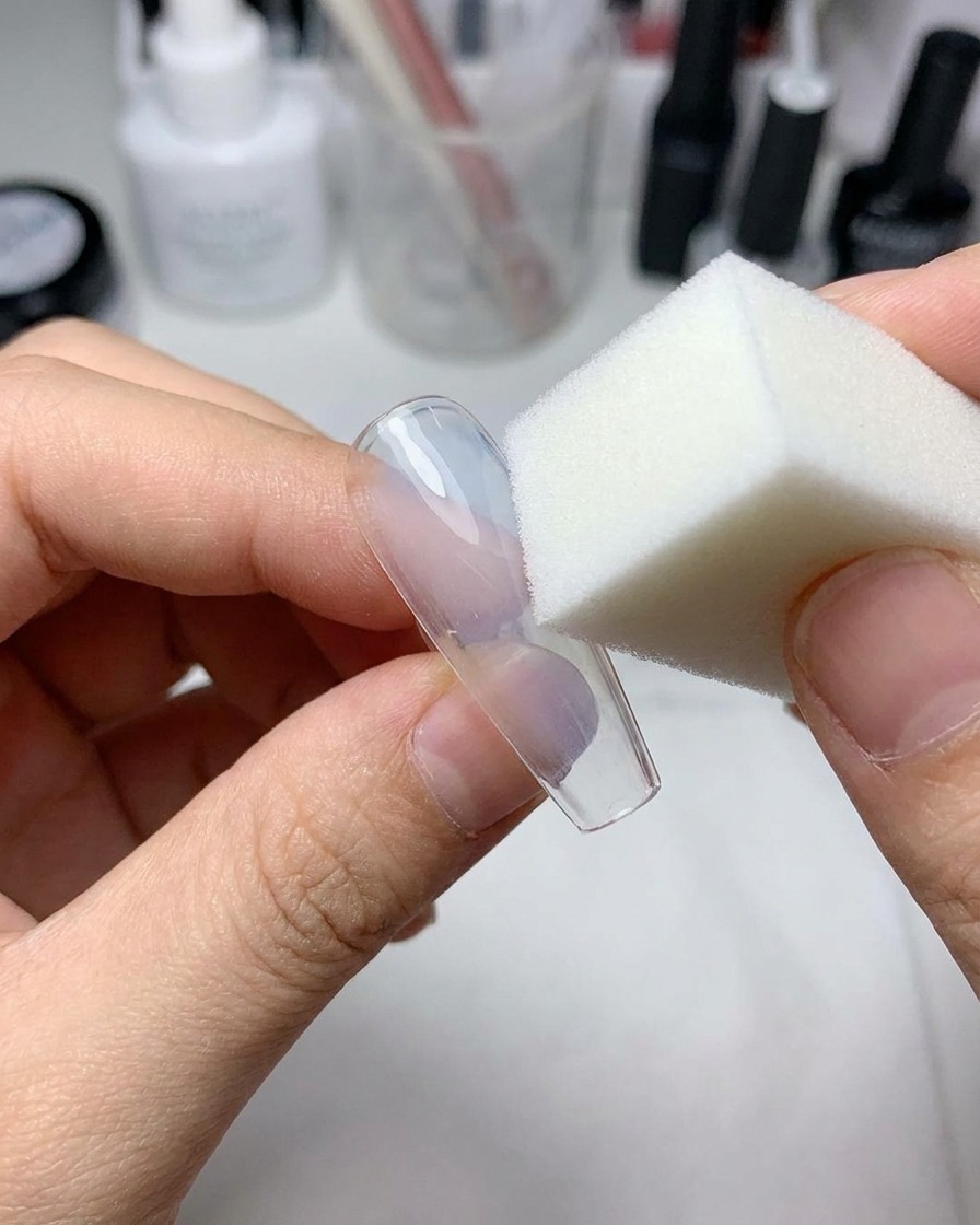 Una piccola spugnetta per trucco che tampona del gel azzurro trasparente al centro di ununghia finta trasparente.