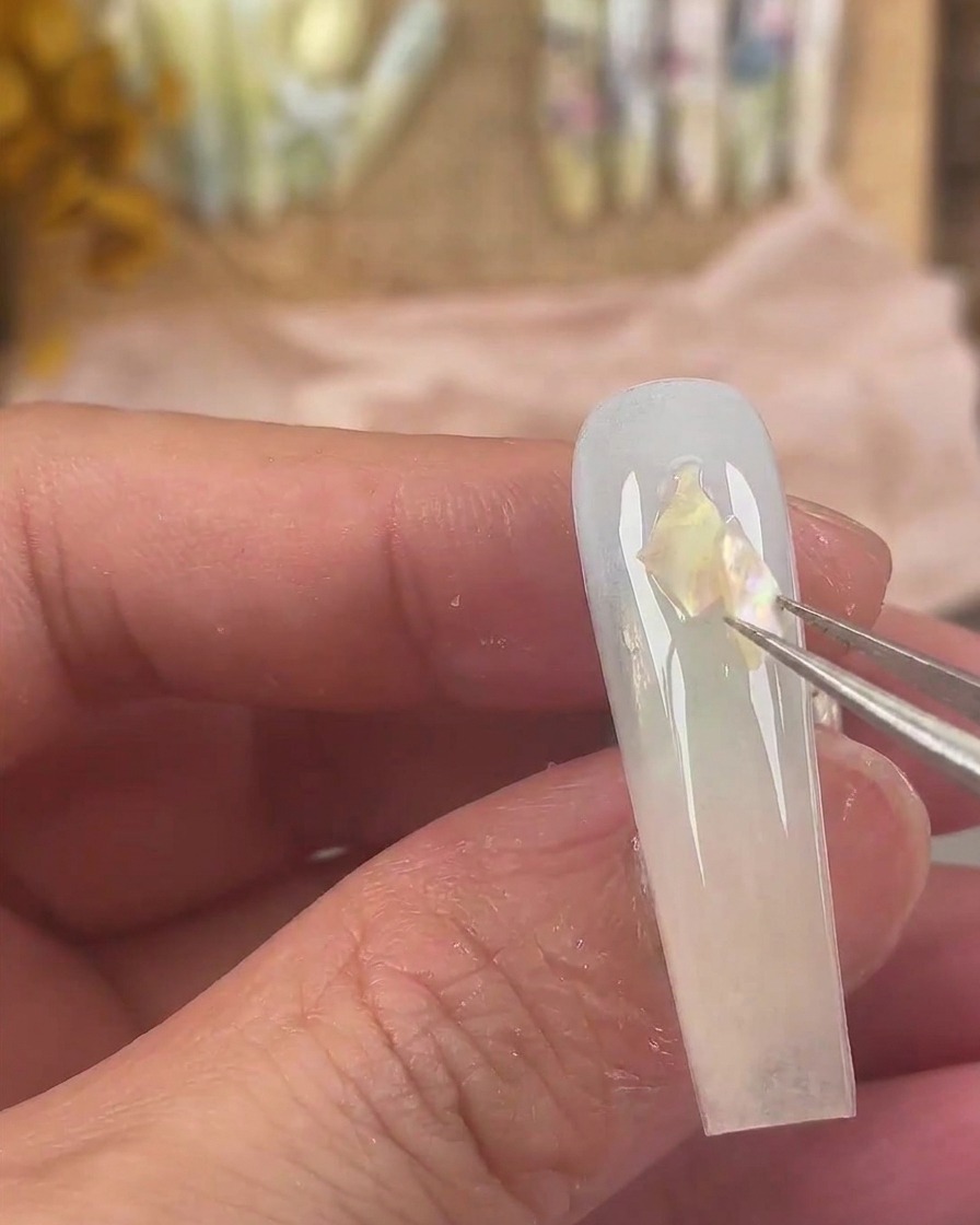 Pinzette a punta fine che posizionano pezzi di conchiglia iridescenti sulla superficie bagnata del gel dellunghia.