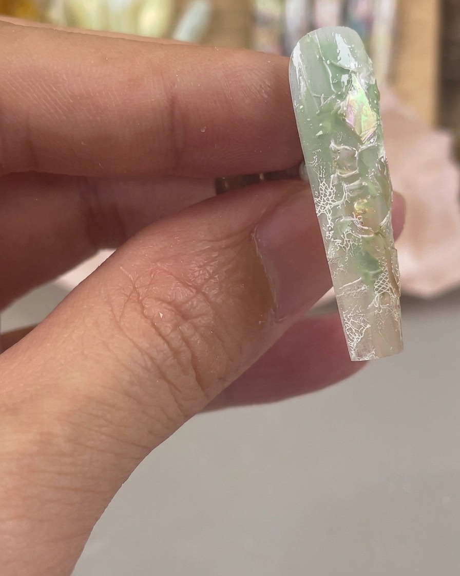 Primo piano di ununghia verde e trasparente con scaglie di conchiglia iridescenti e motivi in pizzo bianco stampati sulla sua superficie.
