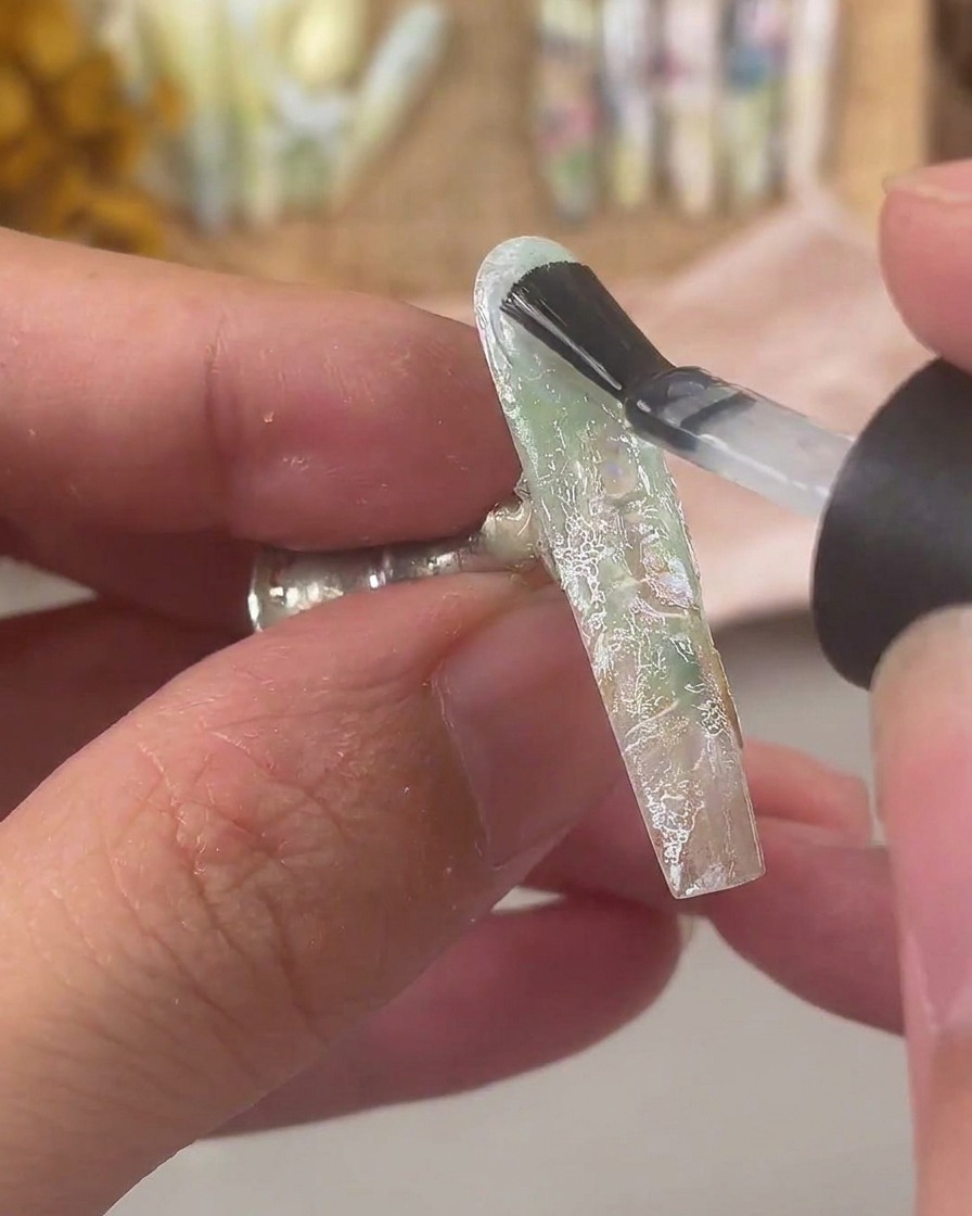 Un pennello da unghie che applica uno strato spesso di builder gel trasparente su ununghia decorata con pizzo e scaglie di conchiglia.