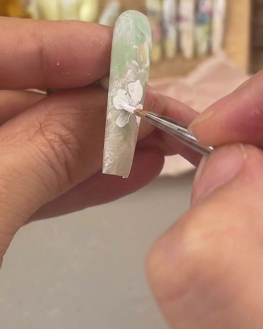 Un pennello da dettaglio fine che applica gel da pittura bianco per disegnare un grande fiore su ununghia verde opaca e trasparente.
