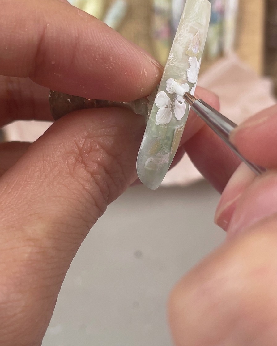 Un pennello da dettaglio fine che dipinge tre piccoli petali floreali bianchi lungo il bordo laterale inferiore di ununghia decorata.