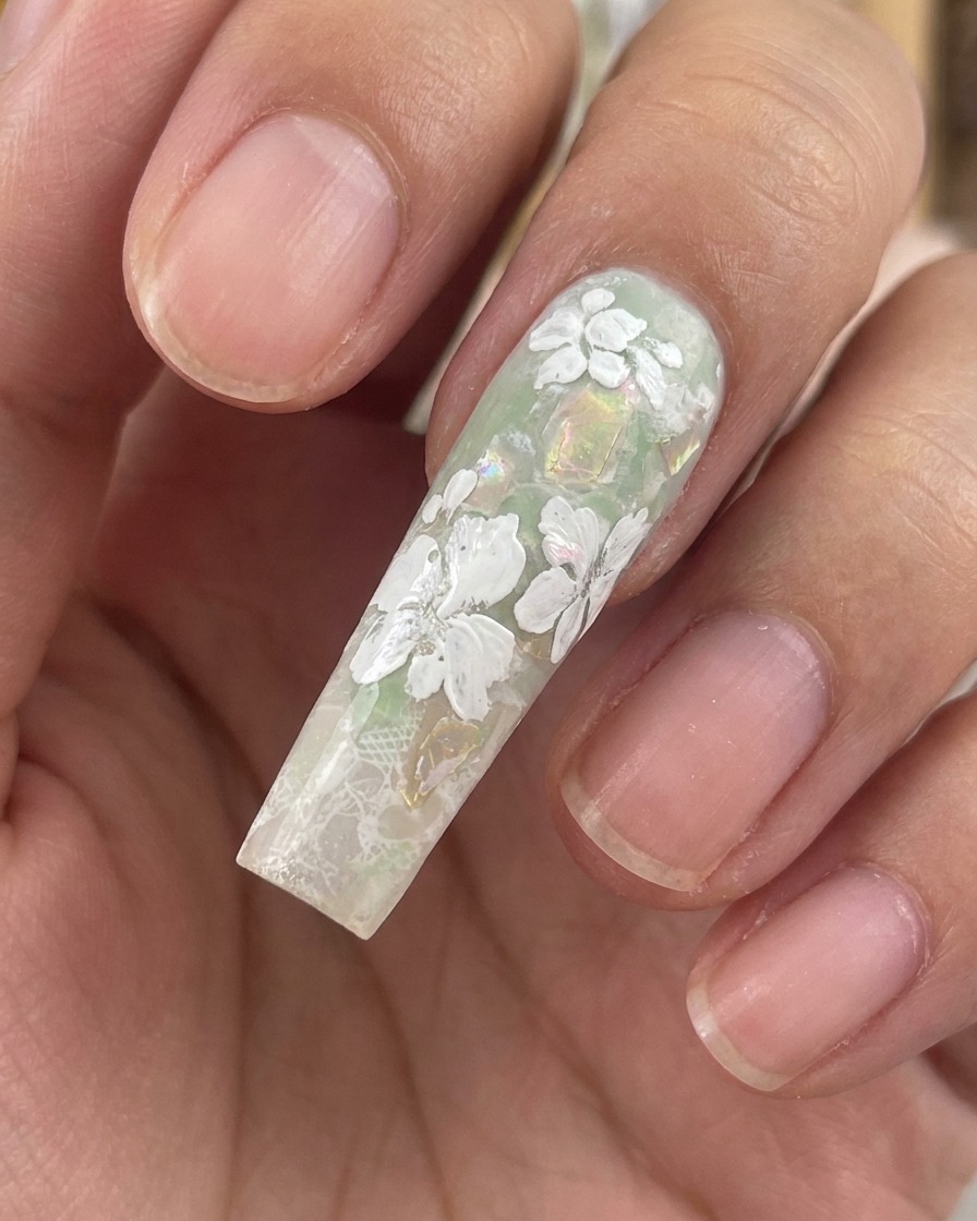 Primo piano di ununghia lunga e scolpita che mostra fiori in gel bianco completati e minuscole foglie su uno sfondo verde e con scaglie di conchiglia.