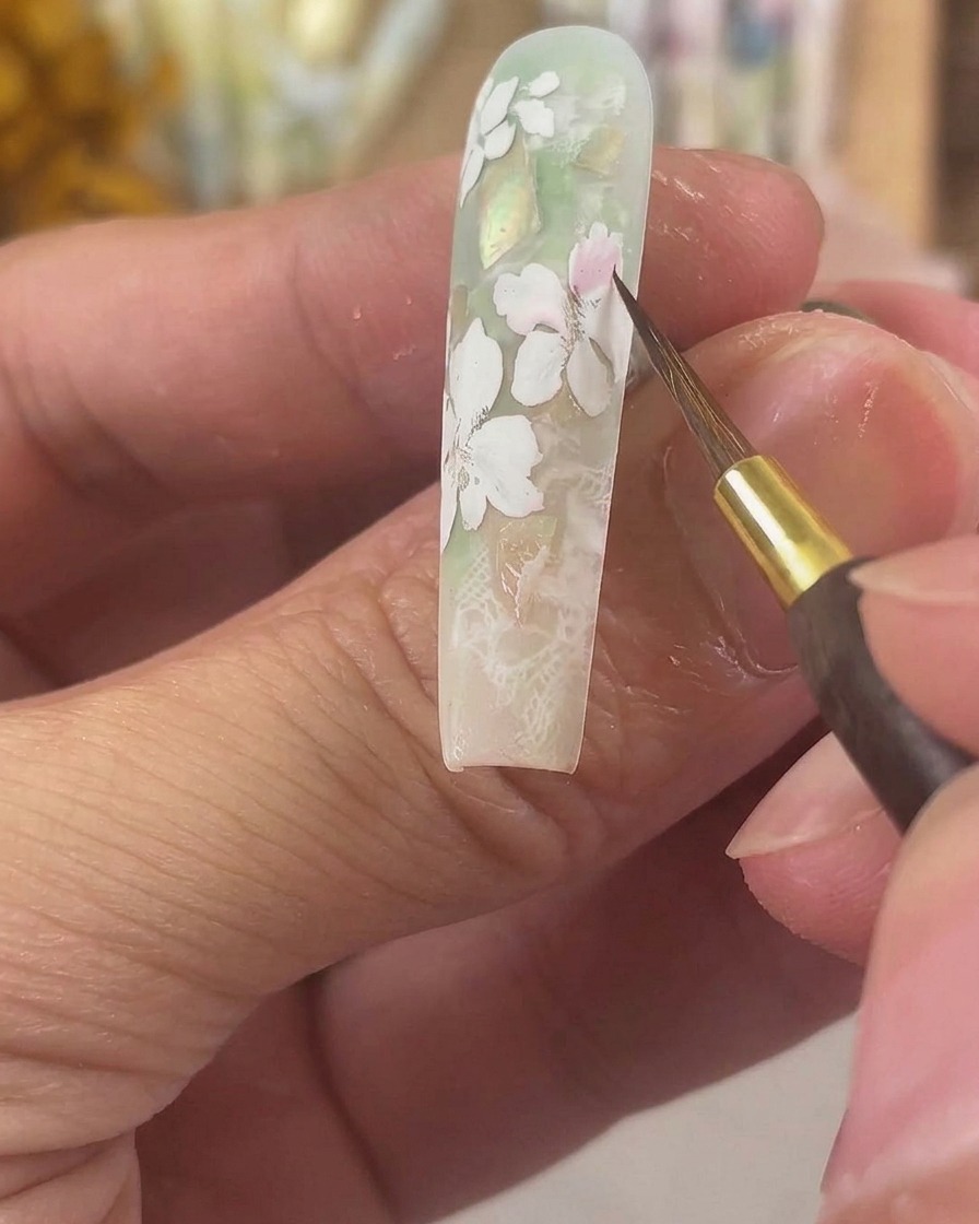 Un pennello da dettaglio fine che applica pigmento acquerello rosa trasparente su petali di gel bianco opaco su ununghia lunga scolpita.