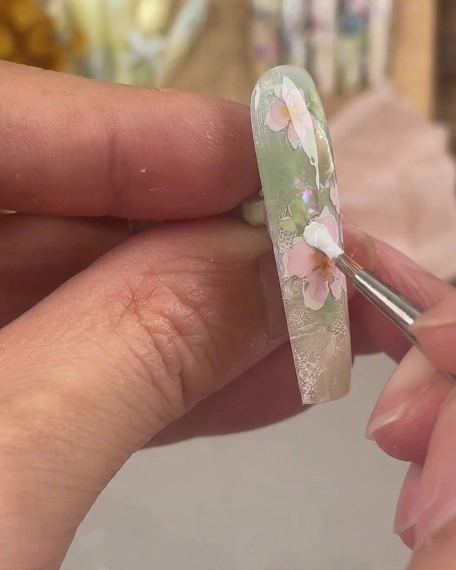 Un pennello liner fine che applica gel da pittura bianco per formare un ulteriore strato di petali su un design di nail art floreale rosa.