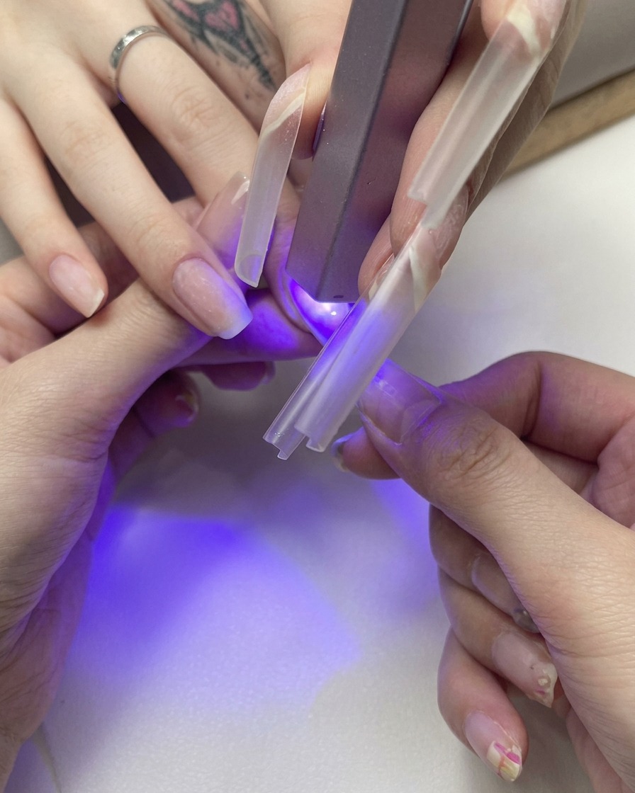 Primo piano di una mano che tiene una tip trasparente per estensione su ununghia naturale mentre polimerizza sotto una piccola lampada UV viola.