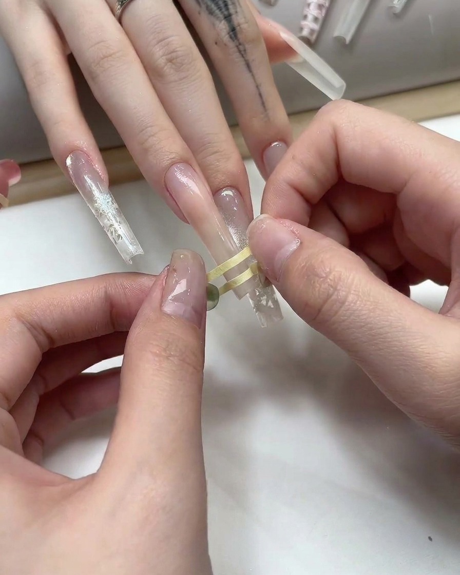 Dita che posizionano del sottile nastro adesivo striping dorato in un motivo a griglia su ununghia in gel nude.