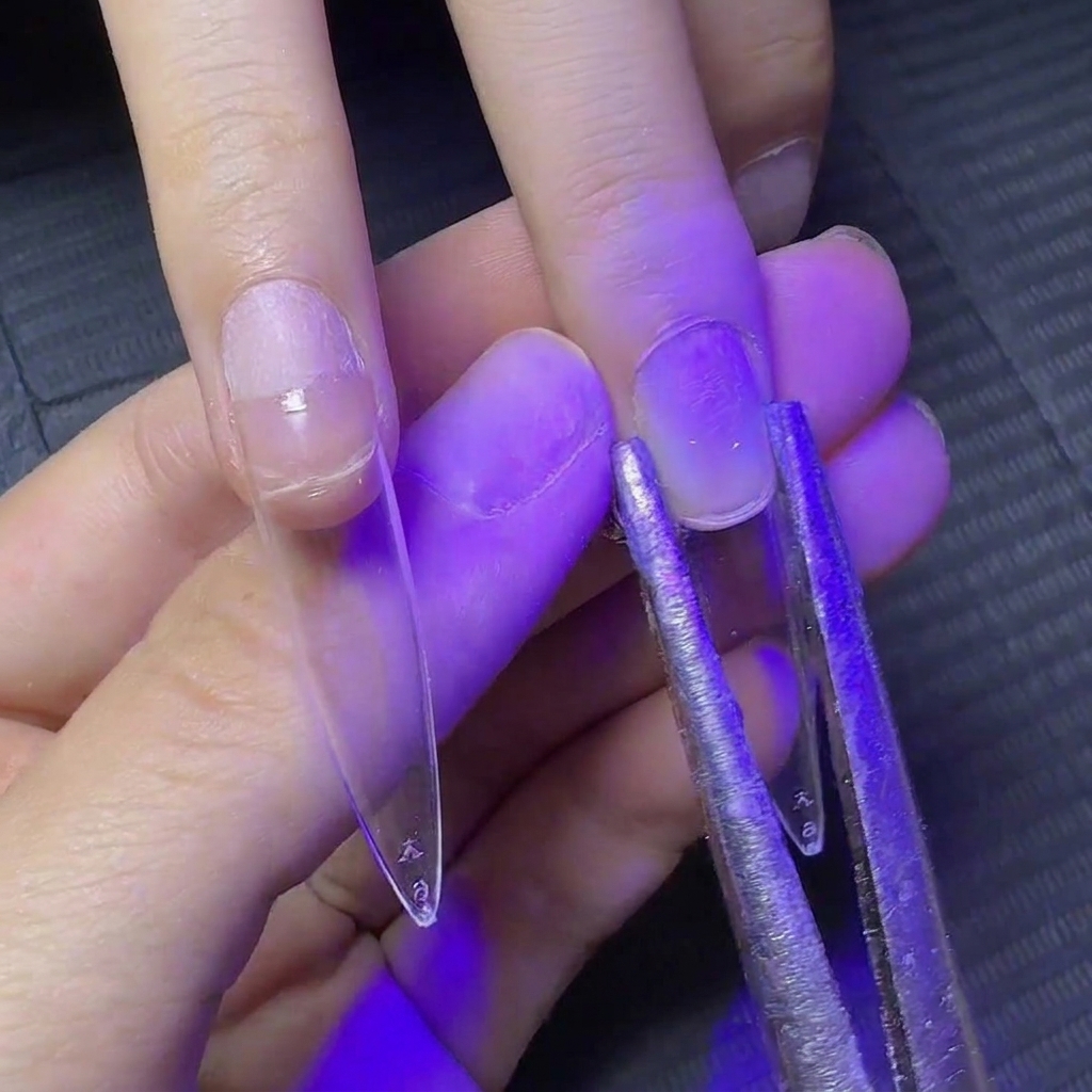 Pinzette metalliche che stringono i lati di ununghia con allungamento trasparente sotto la luce UV per modellarne la curva.