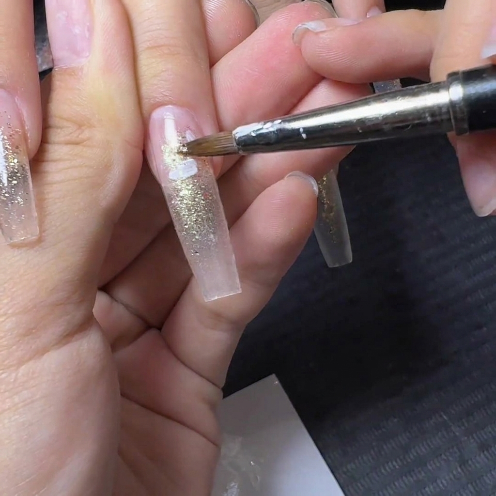 Un pennello da dettaglio fine che applica design di petali di fiori bianchi su ununghia glitterata traslucida.